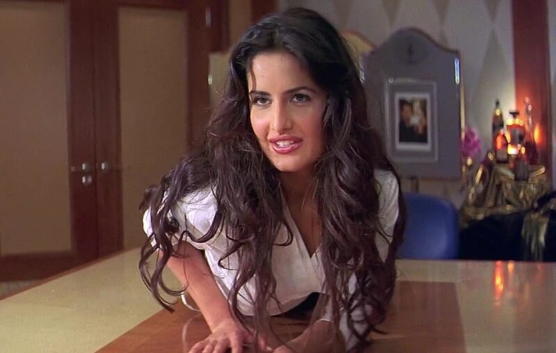 Katrina Kaif with long, wavy hair leans forward on a polished wooden table, smiling playfully in Boom 2003 Film